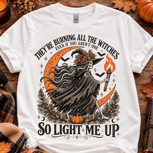 May include: A white short-sleeve t-shirt featuring a graphic of a skeleton witch wearing a hat with autumn leaves, holding a flaming torch. The design includes an orange crescent moon, bats, and the text: THEY'RE BURNING ALL THE WITCHES EVEN IF YOU AREN'T ONE SO LIGHT ME UP.