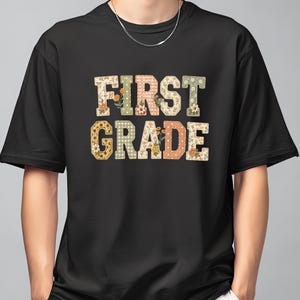 May include: A black short-sleeved t-shirt featuring the text FIRST GRADE in large block letters. Each letter is filled with a different pattern, including gingham, polka dots, and floral prints in muted shades of orange, green, and cream.