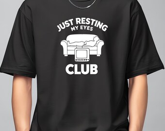May include: Black t-shirt with a white graphic. The graphic features the text "JUST RESTING MY EYES CLUB" above an illustration of a person resting on a couch in front of a television.