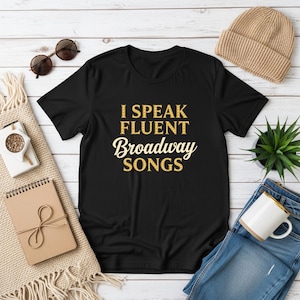 Pode incluir: Camiseta preta com o texto "I Speak Fluent Broadway Songs" em letras douradas com glitter. A camisa é cercada por acessórios, incluindo óculos de sol, um gorro bege, uma pequena planta em vaso, uma caneca de café e um caderno.