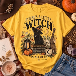 May include: A mustard yellow t-shirt featuring a graphic of a black cat wearing a witch hat sitting on a stack of books labeled Spells, Herbs, and Potions, surrounded by a lantern, a pumpkin, a skull, a broom, and the text There's a Little Witch in All of Us.