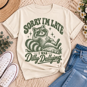Puede incluir: Una camiseta color crema con un gráfico verde oscuro de un mapache con gafas de sol, recostado mientras sostiene una bebida. El texto dice "SORRY I'M LATE" sobre el mapache y "I WAS DILLY DALLYING" debajo, con pequeños detalles de estrellas.