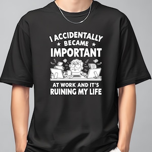 Op de afbeelding: Zwart T-shirt met de witte tekst "I ACCIDENTALLY BECAME IMPORTANT AT WORK AND IT'S RUINING MY LIFE". De afbeelding toont een gestreste persoon aan een bureau met computers en vliegend papier. Het shirt heeft korte mouwen en een ronde hals.