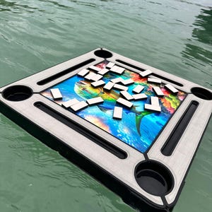 Może przedstawiać: Pływająca plansza do gry w domino z kolorową grafiką ryby pośrodku. Plansza ma szarą ramę z czarnymi uchwytami na kubki i czarną obwódką. Białe kostki domina są rozrzucone po planszy, która unosi się na wodzie.