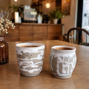May include: Two handmade ceramic coffee mugs with a white and gray marbled design. The mugs are filled with coffee and sit on a wooden table. The mugs are part of a set and are perfect for enjoying a warm beverage.