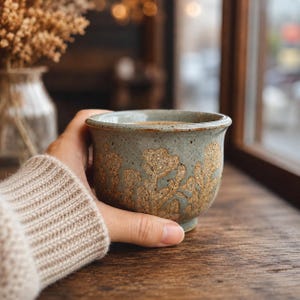 Peut inclure: Une tasse en céramique avec un émail bleu-gris clair moucheté et un motif floral en relief texturé de couleur sable. La tasse présente une forme arrondie à large bord et un rebord légèrement évasé, offrant un aspect artisanal rustique.