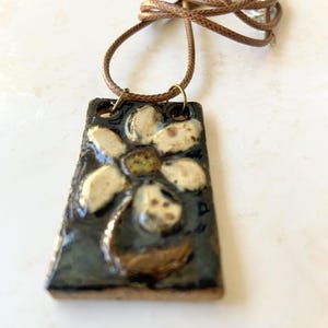 May include: A rectangular ceramic pendant necklace with a floral design. The pendant features a dark background with a raised, cream-colored flower and a brown leather cord. The necklace is a handmade piece.