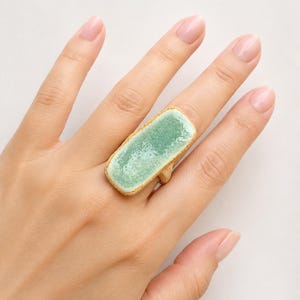 May include: A rectangular ring featuring a textured, light brown base and a smooth, glossy, seafoam green glazed top surface. The ring has an elongated, organic shape with uneven edges, showcasing a contrast between the matte, earthy band and the vibrant, reflective green center.