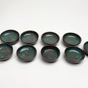 May include: A set of nine small, round ceramic bowls arranged in a grid. Each bowl features a deep teal and green mottled glaze with a dark brown rim and base, showcasing a smooth, glossy finish and a shallow, concave interior design.