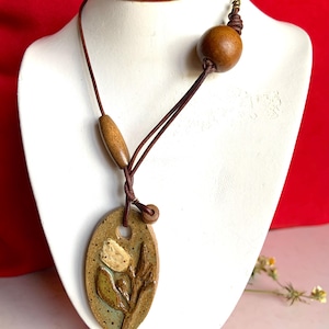 May include: A handcrafted ceramic pendant necklace featuring a floral design. The pendant is oval-shaped with a raised relief of a flower and leaves, in shades of beige, green, and brown. The necklace has a brown leather cord with wooden beads.