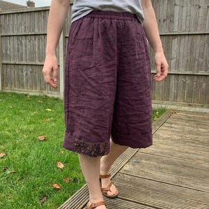 Pode incluir: Culotes de linho cor de ameixa com perna larga e cós elástico. Os culotes têm uma faixa decorativa na bainha e são combinados com sandálias castanhas. A pessoa está vestindo uma camiseta cinza.