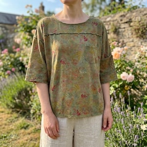 May include: A short-sleeved olive green t-shirt featuring a subtle, multi-coloured floral and botanical print. The garment has a round neckline, a single chest pocket on the left side, and visible horizontal seam detailing across the upper chest.