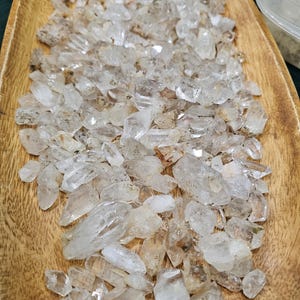 May include: A wooden tray filled with clear quartz crystal points. The crystals vary in size and shape, with some having a slightly cloudy appearance. The tray is made of light brown wood and has a natural, organic shape. The crystals are a popular choice for jewelry and decor.
