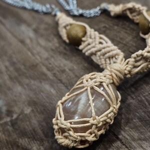 May include: A handmade macrame necklace featuring a large, polished, light brown stone pendant. The pendant is encased in a beige macrame net, with a matching macrame cord and silver chain. The necklace has a natural, bohemian aesthetic.