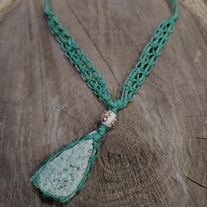 Puede incluir: Collar de macramé hecho a mano con un cordón tejido verde claro. El collar tiene un colgante con una piedra verde claro envuelta en el mismo cordón. Una pequeña cuenta estampada está colocada encima del colgante. El collar se muestra sobre una superficie de madera.