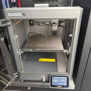 May include: A grey ELEGOO 3D printer with a rectangular build plate and a small touchscreen display. The printer has a closed-frame design and is set on a metal cabinet. The ELEGOO logo is visible on the top and the display.