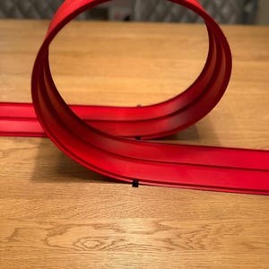 May include: A red plastic toy race track loop-de-loop, designed for toy cars. The track is a vibrant red color and is set against a light brown wooden surface. The loop is a complete circle, adding a fun element to the toy.
