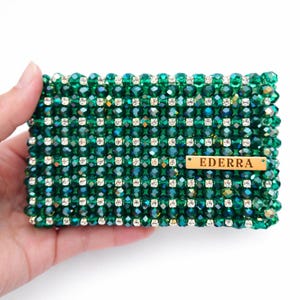 May include: A rectangular, beaded clutch in emerald green, adorned with rows of sparkling clear crystals. The clutch features a small, rectangular wooden tag with the name "EDERRA" engraved on it. The beads are arranged in a grid pattern.
