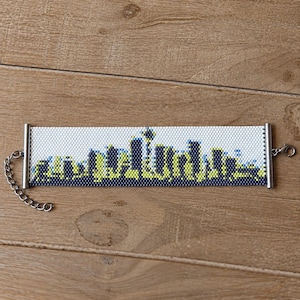 May include: A wide beaded bracelet featuring a pixelated cityscape design in shades of white, navy blue, and lime green. The bracelet has silver-toned metal end bars, a lobster claw clasp, and an adjustable chain link extension on one side.