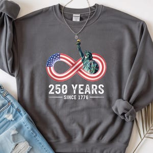 May include: Dark gray sweatshirt with the text "250 YEARS SINCE 1776" and an infinity symbol design featuring the American flag and the Statue of Liberty. A silver necklace and blue jeans are also in the image.