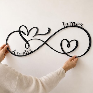 May include: A black wall-mounted sign in the shape of an infinity symbol, featuring the names Amelia and James integrated into the design. The sign includes two stylised heart shapes incorporated into the loops of the infinity symbol.