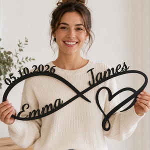 May include: A black, laser-cut wall sign in the shape of an infinity symbol. The sign features the name Emma, the name James, a heart shape, and the date 05.10.2026 integrated into the continuous, flowing lines of the infinity design.
