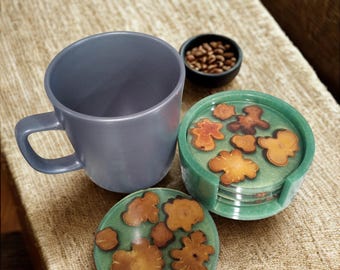 Puede incluir: Una taza de cerámica gris con asa se encuentra junto a un juego de posavasos de resina verde. Los posavasos presentan un diseño de formas de madera marrón incrustadas en la resina. También hay un pequeño cuenco negro con granos de café.