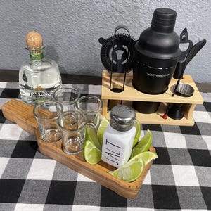 May include: A wooden serving tray holding four clear glass shot glasses, a white salt shaker with the word SALT printed on it, and several lime wedges. Next to it is a wooden stand with a black cocktail shaker and bar tools.