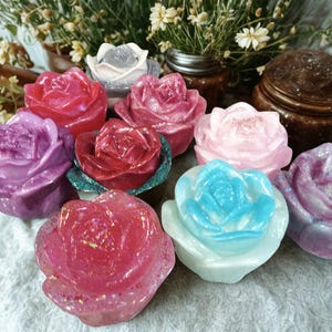 Może przedstawiać: Kolekcja kolorowych, dekoracyjnych przedmiotów w kształcie róży. Róże są w różnych odcieniach różu, fioletu, czerwieni i niebieskiego, z brokatowymi akcentami. Przedmioty ułożone na białej powierzchni, z zielenią w tle.