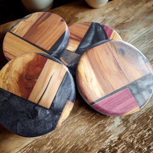 May include: Four round wooden coasters with a geometric design. The coasters feature a combination of light and dark wood tones, with accents of black resin and a deep red hue. The coasters are arranged on a wooden surface, with two white mugs in the background.