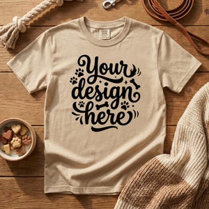 May include: Tan t-shirt with the text "Your design here" in black script. The shirt is surrounded by a dog leash, rope toy, dog treats, and a sweater. The items are on a wooden surface.