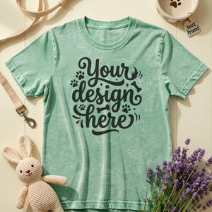 May include: A light green t-shirt with the text "Your design here" in black script, paw prints, and a bone graphic. The image includes a dog bowl, a leash, a stuffed bunny, and lavender.