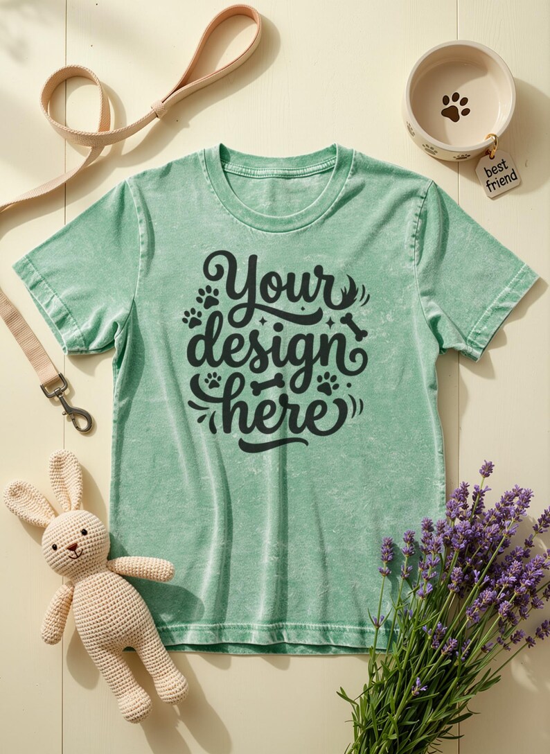 May include: A light green t-shirt with the text "Your design here" in black script, paw prints, and a bone graphic. The image includes a dog bowl, a leash, a stuffed bunny, and lavender.