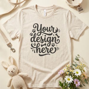 May include: Beige t-shirt with the text "Your design here" in a black, decorative font. The image includes a dog bowl, a leash, a stuffed bunny, and a bouquet of flowers. The dog bowl has a paw print.