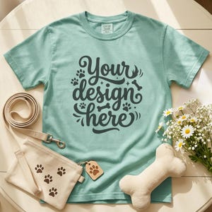 May include: A light teal t-shirt with the text "Your design here" in a dark gray script. A beige dog leash, a small beige bag with paw prints, a wooden tag, a plush bone, and a bouquet of daisies are also present.