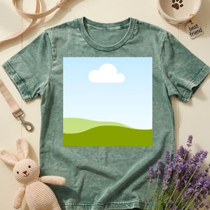 May include: A green t-shirt with a landscape graphic featuring a blue sky, white cloud, and green hills. The image also includes a beige dog leash, a ceramic pet bowl with a paw print, a stuffed bunny, and lavender sprigs.