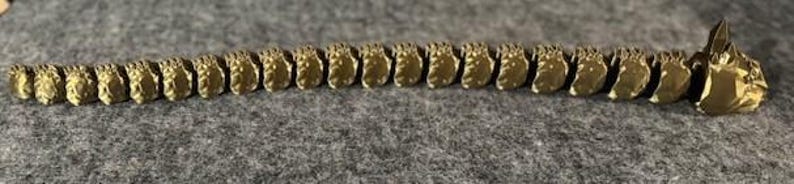 May include: A gold-coloured, segmented spine-shaped object, possibly a decorative piece or jewellery component. The segments are detailed and arranged linearly, with a textured surface. The object is lying on a grey, textured surface.