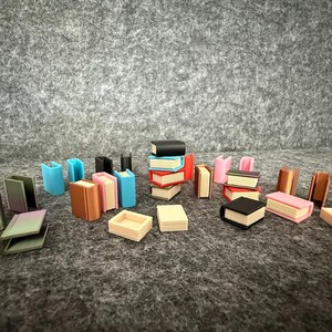 Può includere: Una collezione di libri in miniatura e oggetti a forma di libro in vari colori, tra cui nero, rosso, rosa e blu. Alcuni sono impilati, altri sono aperti o in piedi. Gli oggetti sono disposti su una superficie grigia.