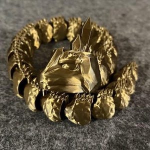 May include: A gold-coloured, 3D-printed decorative ring with a detailed, textured design. The ring features a central structure with sharp, crystalline elements and a surrounding band of repeating, organic shapes. The overall aesthetic is ornate and fantasy-inspired.