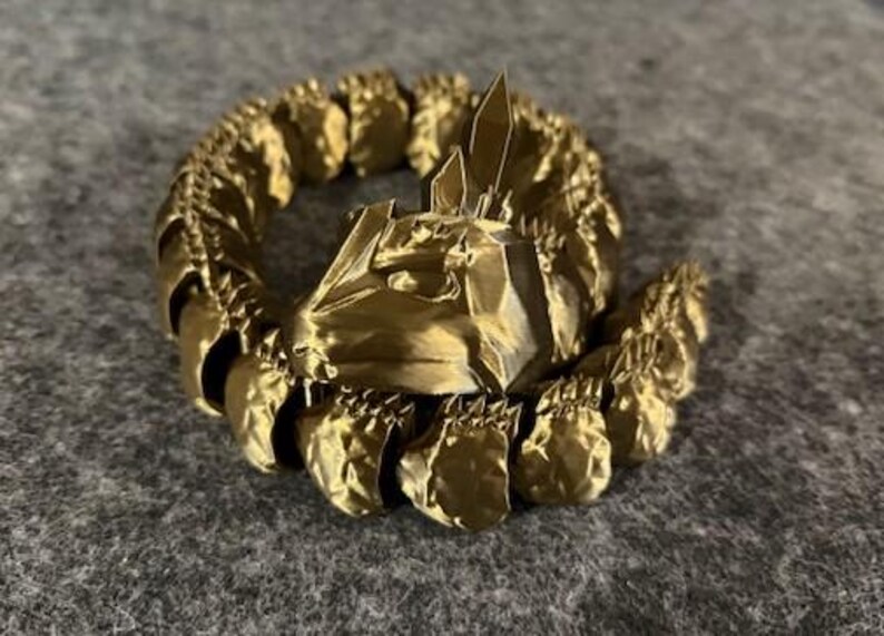 May include: A gold-coloured, 3D-printed decorative ring with a detailed, textured design. The ring features a central structure with sharp, crystalline elements and a surrounding band of repeating, organic shapes. The overall aesthetic is ornate and fantasy-inspired.