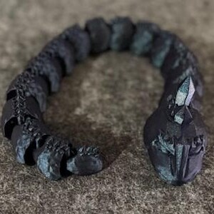 May include: A dark grey articulated snake figurine with a faceted crystal on its head. The snake has a textured, segmented body with hints of teal. The figurine is posed in a curved shape.