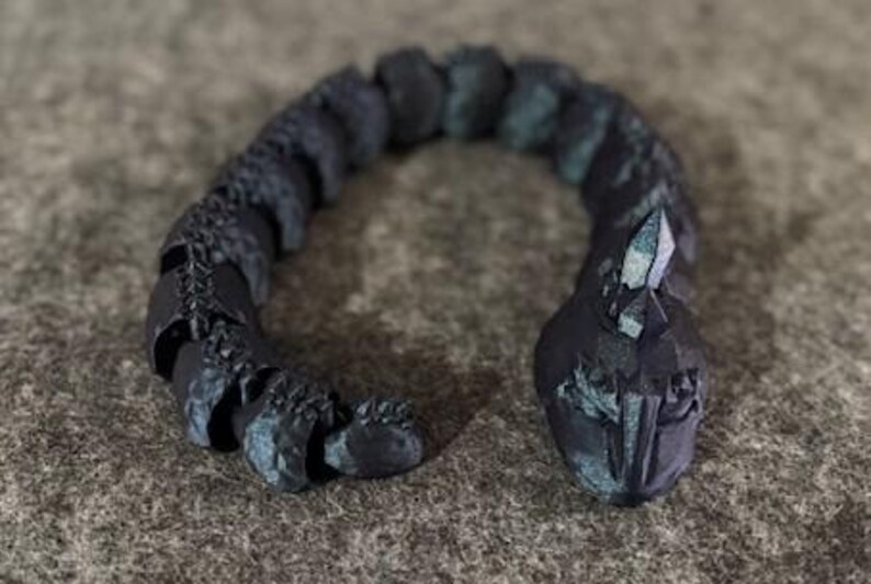 May include: A dark grey articulated snake figurine with a faceted crystal on its head. The snake has a textured, segmented body with hints of teal. The figurine is posed in a curved shape.