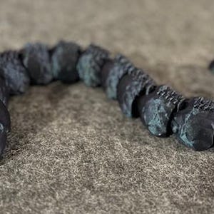 May include: A dark, segmented sculpture of a serpentine creature with a crystalline head. The body is composed of connected, textured segments in shades of blue and grey. The sculpture is displayed on a neutral, textured surface.