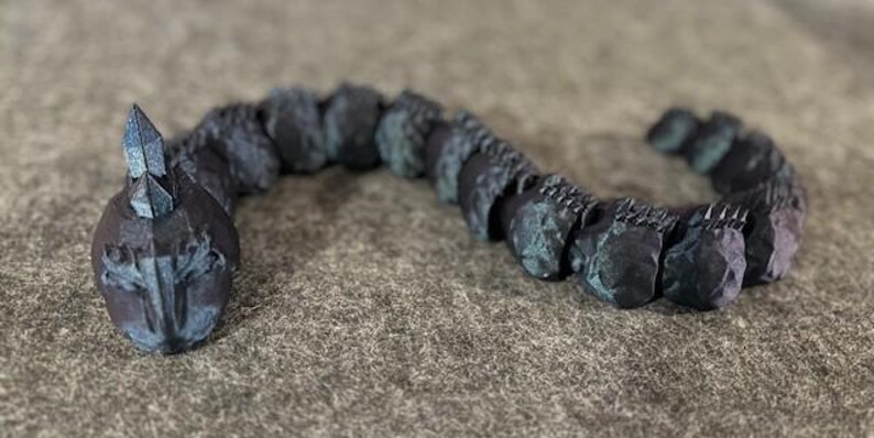 May include: A dark, segmented sculpture of a serpentine creature with a crystalline head. The body is composed of connected, textured segments in shades of blue and grey. The sculpture is displayed on a neutral, textured surface.