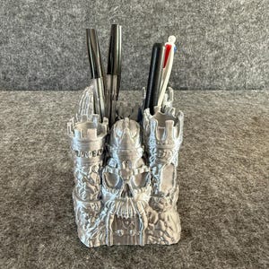 May include: A silver-colored pen holder shaped like a castle with a skull design. The pen holder has three sections and is filled with pens. The castle design includes turrets and a detailed facade.