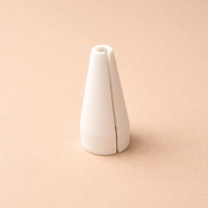 Könnte beinhalten: Ein weißes, kegelförmiges Objekt mit einem vertikalen Schlitz an der Seite. Das Objekt hat eine abgerundete Basis und eine zylindrische Oberseite. Der Hintergrund ist einfarbig hellpfirsichfarben. Das Objekt scheint aus Keramik oder einem ähnlichen Material zu bestehen.