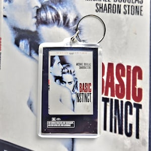 May include: A rectangular clear acrylic keychain featuring a miniature replica of the Basic Instinct movie poster. The insert displays Michael Douglas and Sharon Stone, the title Basic Instinct in red, and an R rating box at the bottom.