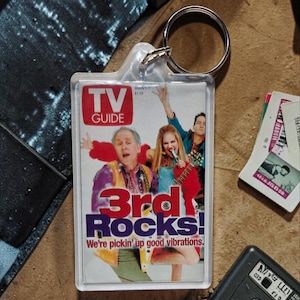 Könnte beinhalten: Ein rechteckiger Schlüsselanhänger aus klarem Acryl mit einem TV Guide-Cover-Motiv. Das Bild zeigt drei Personen mit dem Text TV GUIDE, 3rd Rocks! und We're pickin' up good vibrations. Der Schlüsselanhänger hat einen silbernen Metallring.