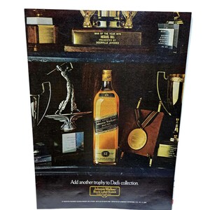 Puede incluir: Anuncio con una botella de whisky Johnnie Walker Black Label Scotch, rodeada de trofeos. El texto dice "A&ntilde;ade otro trofeo a la colecci&oacute;n de pap&aacute;" y "Hombre del A&ntilde;o 1976".