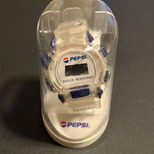 May include: A Pepsi-branded digital watch in a clear plastic case. The watch face is white with a black digital display and the Pepsi logo. The watch has blue accents and the words "SHOCK RESISTANT" are visible.